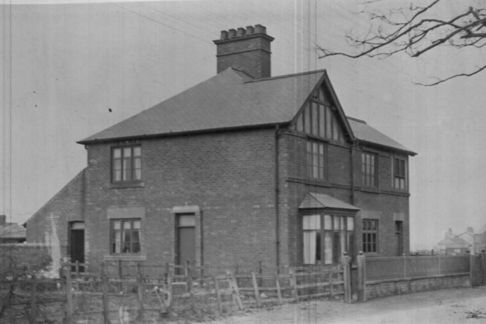 Black-and-white photo of a two-story brick house with multiple chimneys, a gabled roof, and fenced front yard. Bare tree branches and a gate are visible in the foreground.
